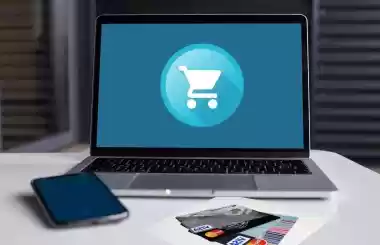 Laptop showing an online shopping cart icon with a phone and bank cards on a desk